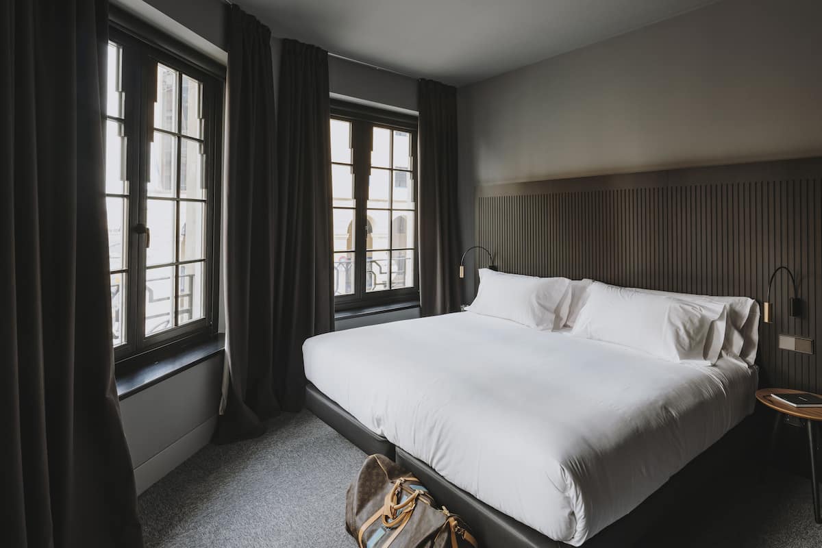 Ein Hotelzimmer mit einem großen Bett, dunklen Vorhängen und Blick durch die Fenster.