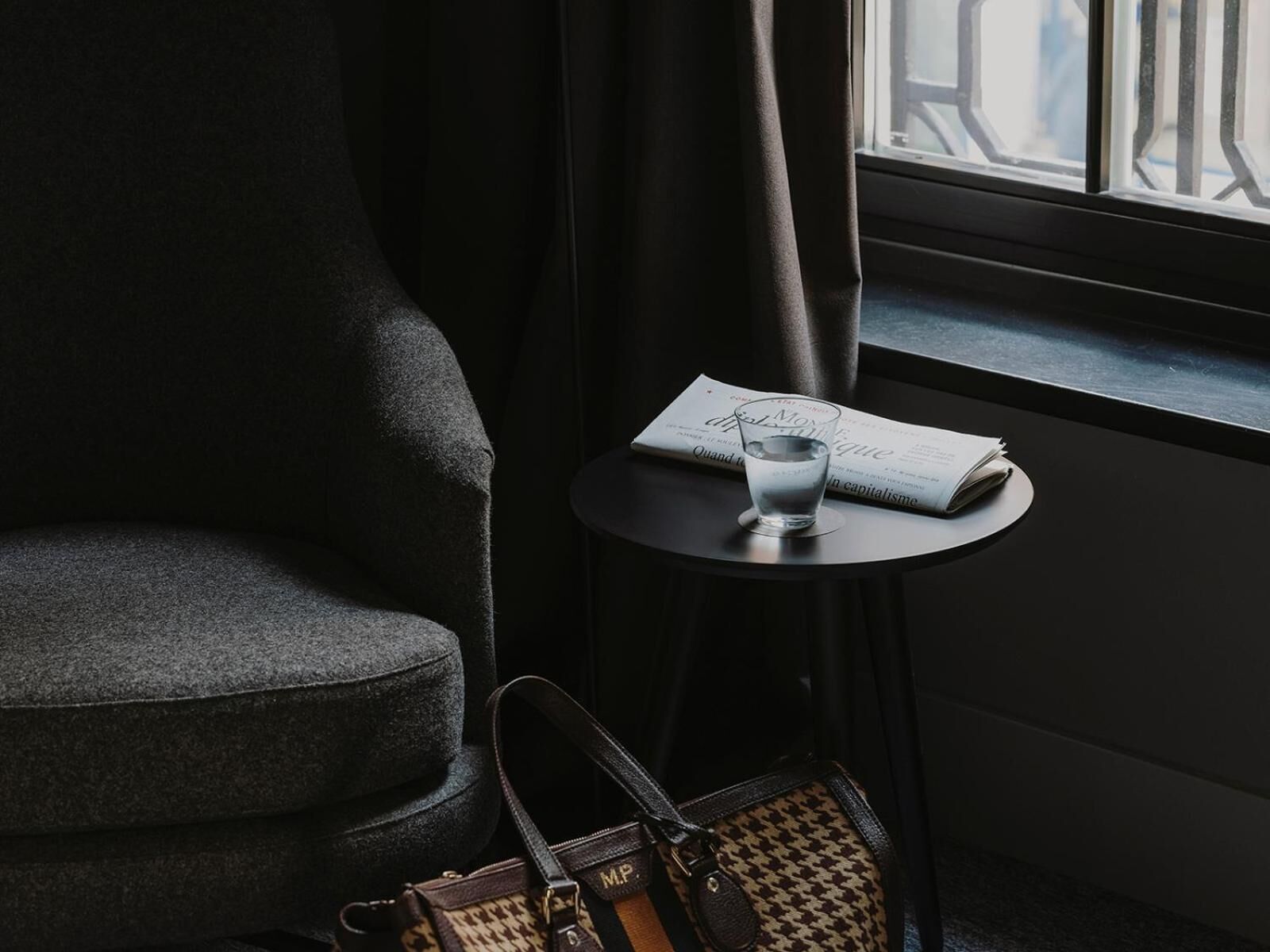Un sillón oscuro, una mesita redonda pequeña con un vaso de agua y una revista, y un bolso de mano.