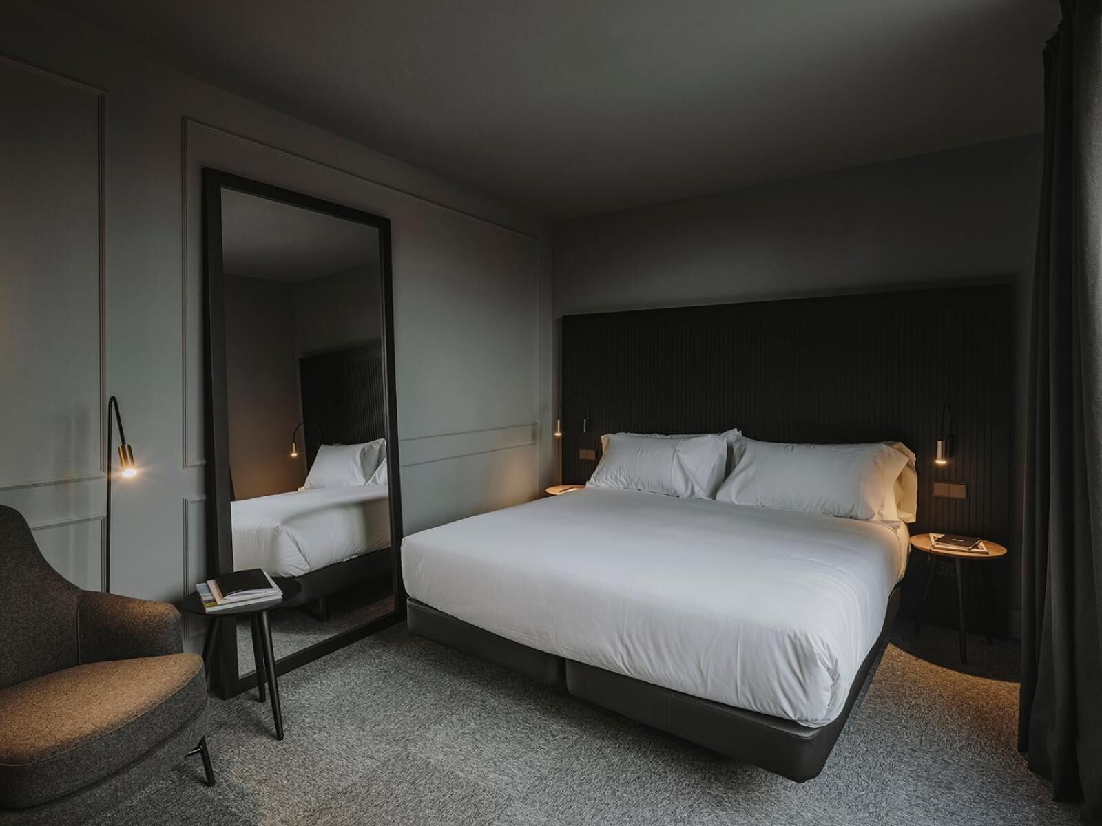 A modern hotel room with a large bed, a seating area with a chair, a small table, and a large mirror.