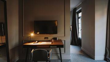 A modern hotel room with a flat-screen TV mounted on the wall, a wooden desk with a chair, and a large window providing natural light.