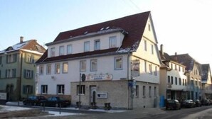 A two-story building with a red-tiled roof, a signboard, and parked cars in front. - Hotel Filoxenia (Stuttgart)