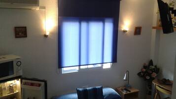 Un dormitorio con cama, mesita de noche, escritorio, lámpara, refrigerador y ventana con persianas.