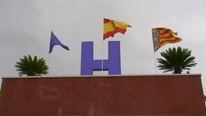 A building with a large blue 'H' and two flags on top. - Hostal Arneva (Orihuela)