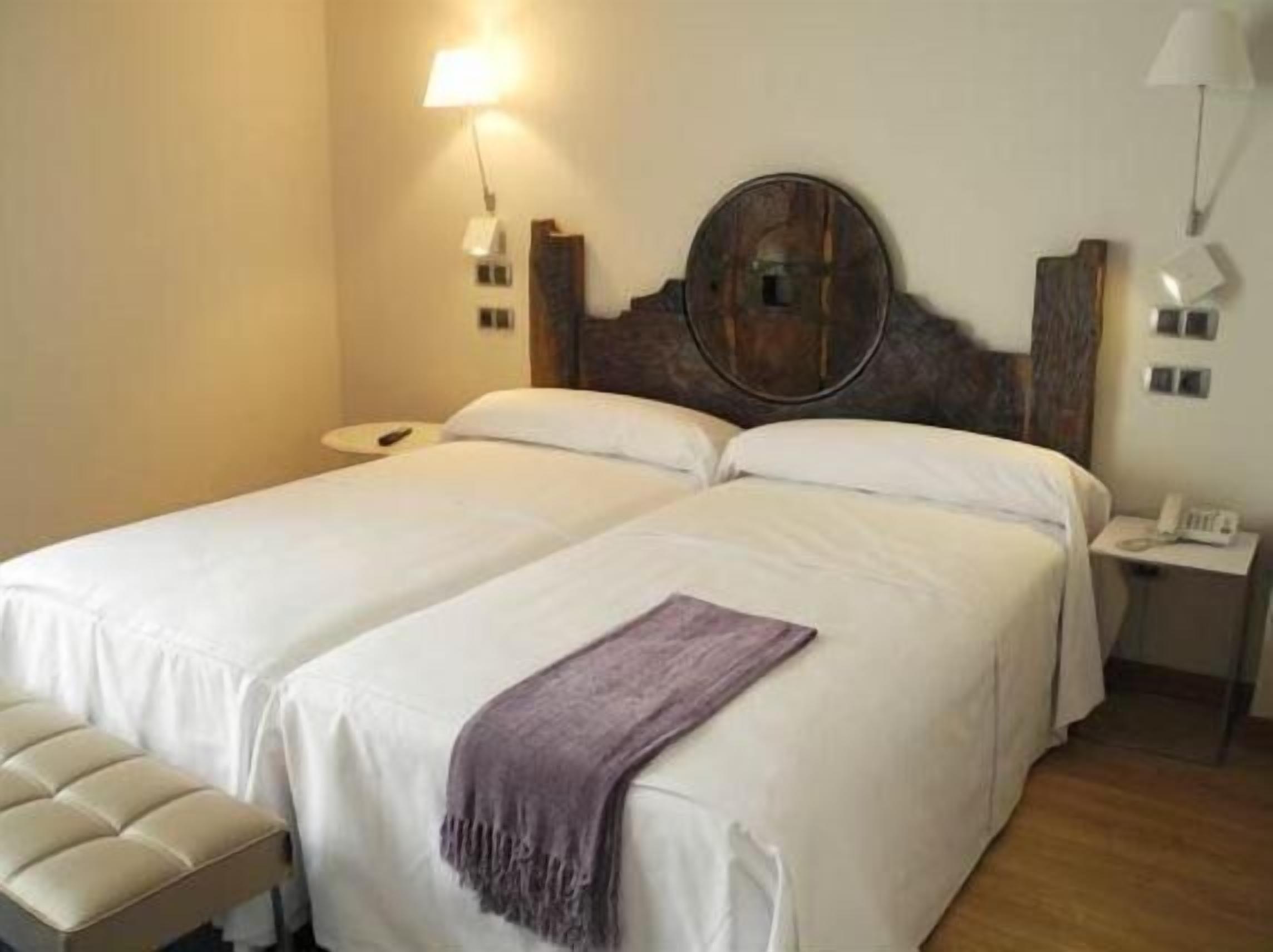 Habitación de hotel con dos camas, una mesita de noche con lámpara, teléfono y un espejo redondo en la cabecera.