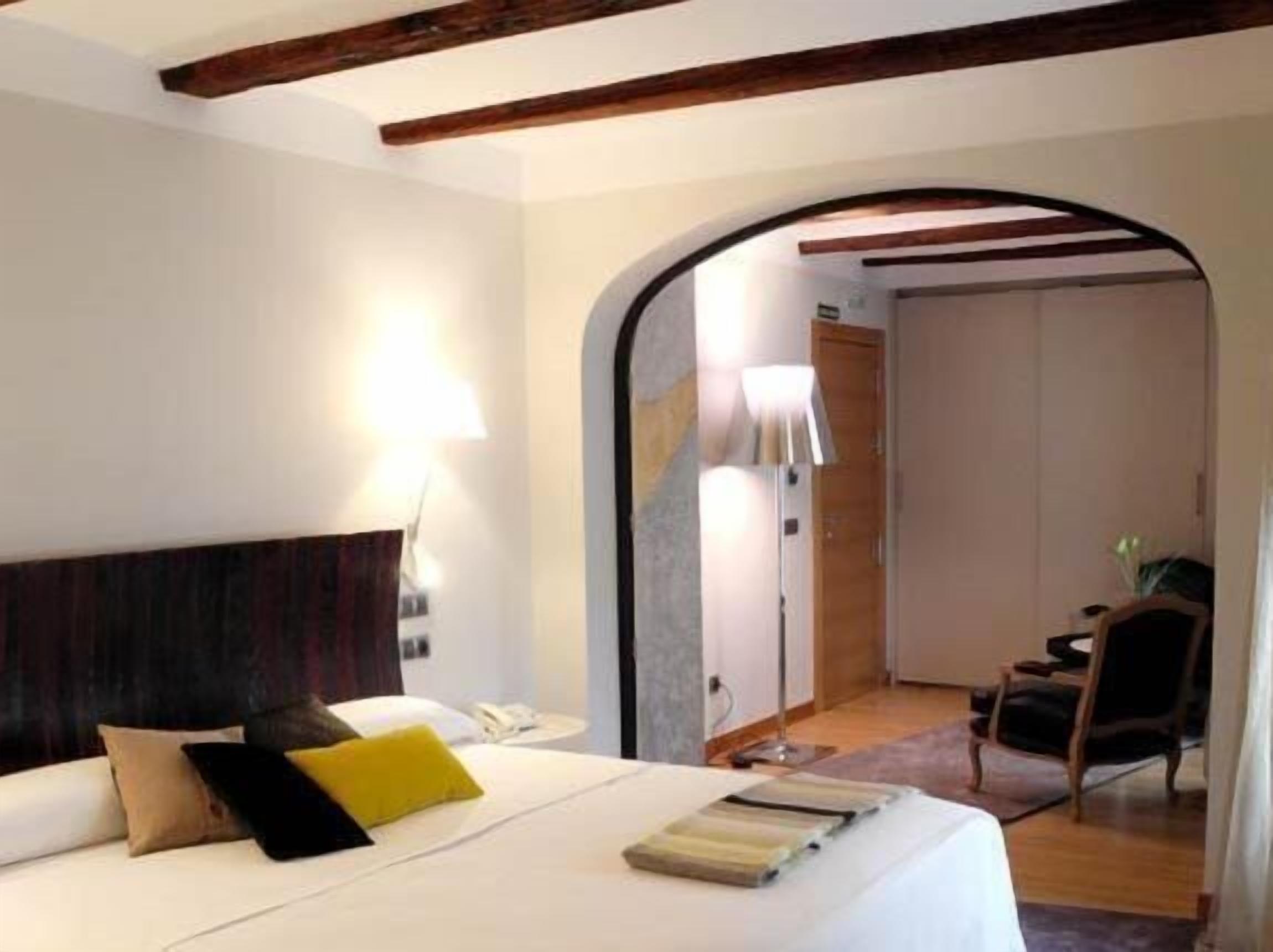 A hotel room with a bed, a desk, and a chair. There is a lamp on the desk and a door leading to another room.