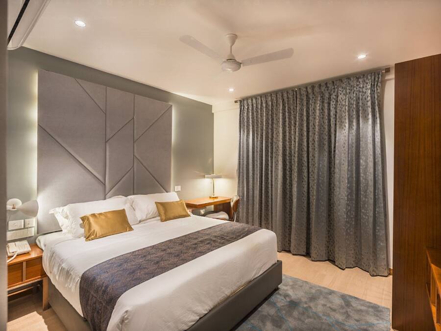 Una habitación de hotel moderna con una cama grande, un ventilador de techo, una mesita de noche con lámpara y una ventana con cortinas.