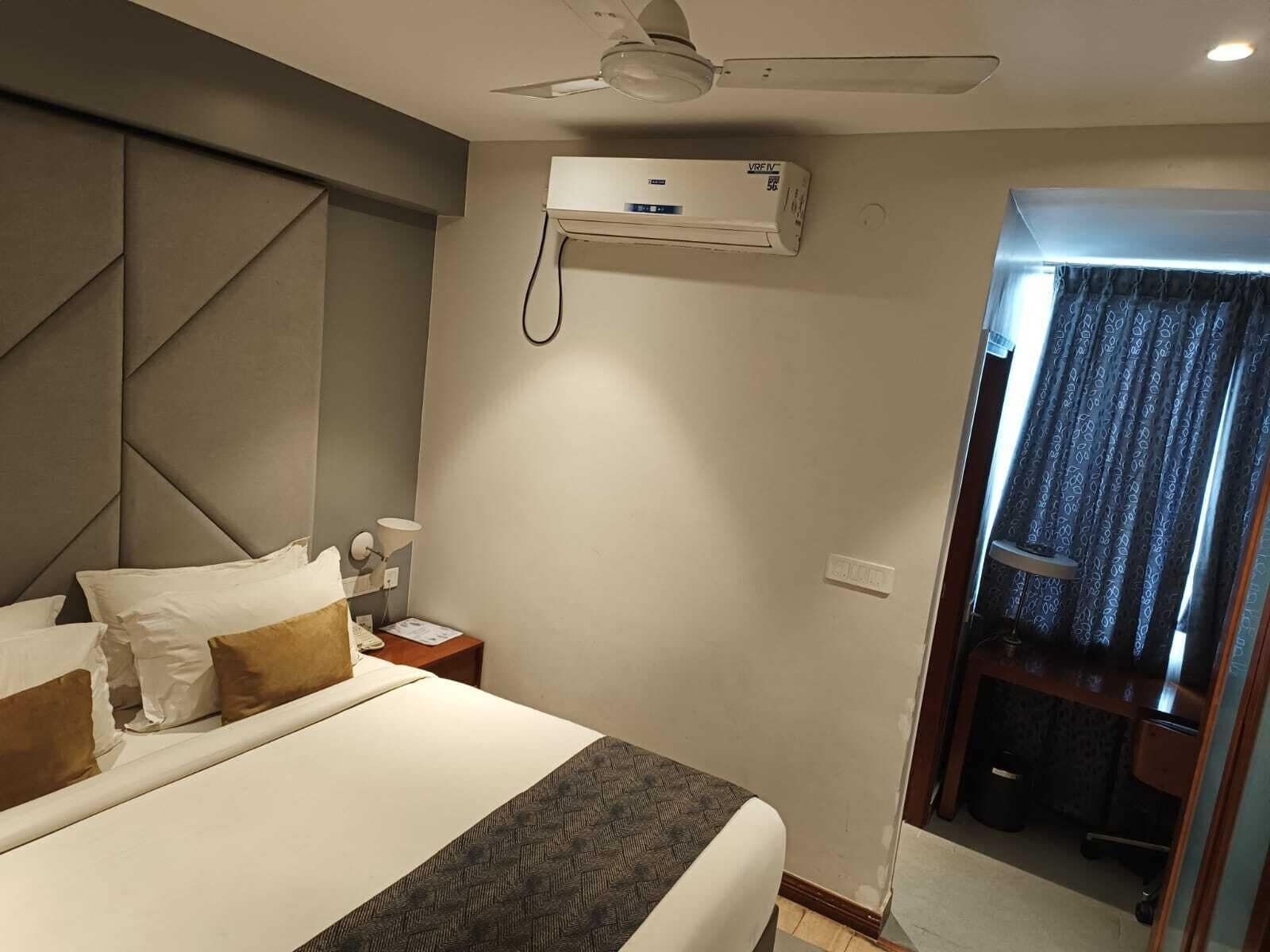A hotel room with a large bed, a ceiling fan, an air conditioning unit, a bedside table with a lamp, and a view of a balcony with curtains.