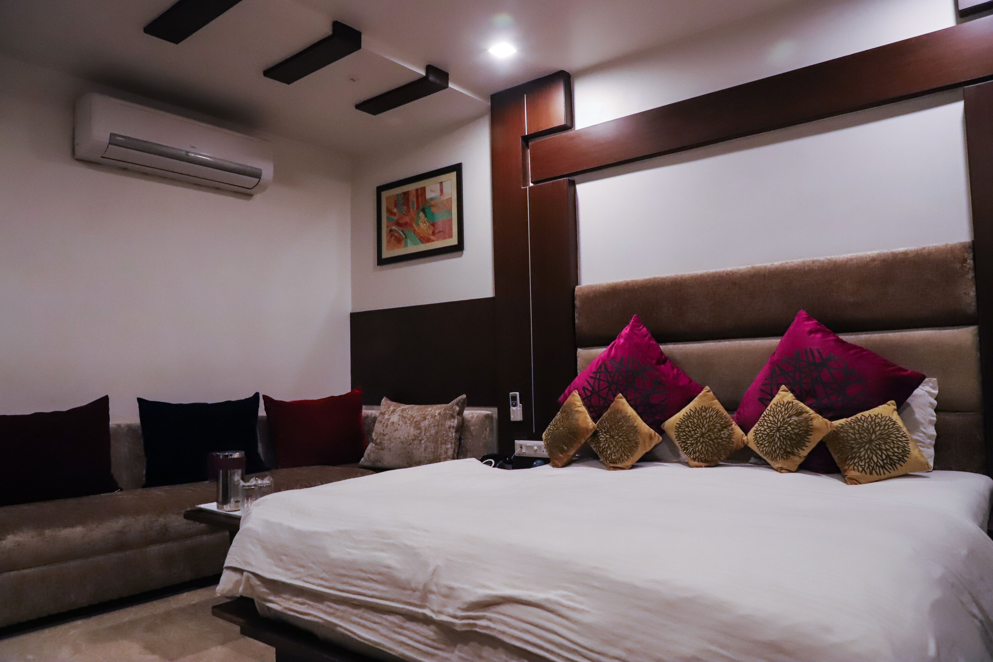 A hotel room with a large bed, a sofa, a coffee table, and an air conditioning unit on the wall.