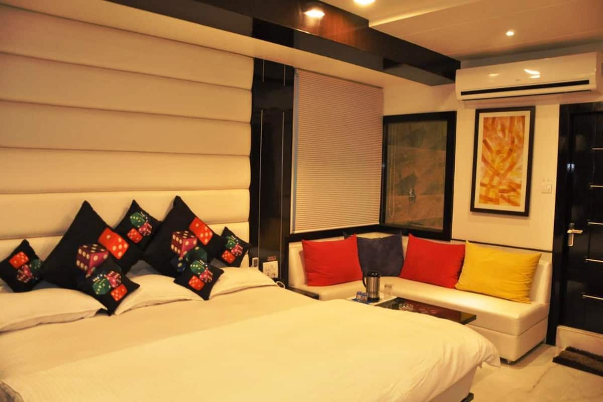 Une chambre d’hôtel moderne avec un grand lit, un canapé et une table basse. On y trouve une fenêtre avec des stores, un tableau au mur et un appareil de climatisation.
