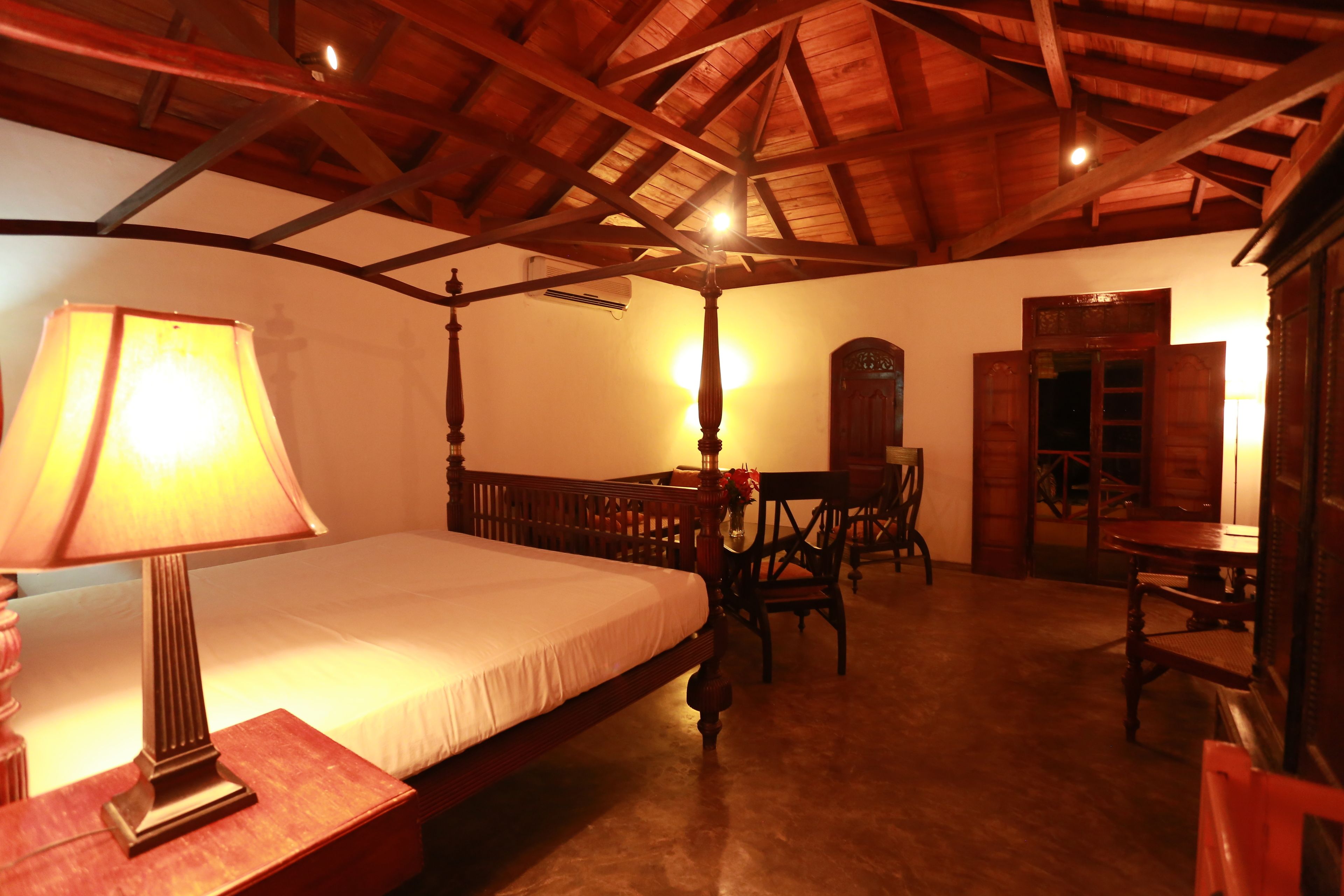 A room with a bed, a table, and a chair. There is a lamp on the table. The room has wooden beams on the ceiling.
