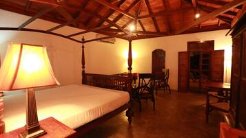A room with a bed, a table, and a chair. There is a lamp on the table. The room has wooden beams on the ceiling.