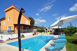 Een zwembad met ligbedden en een grote parasol, een gebouw met balkon, en een helderblauwe lucht.