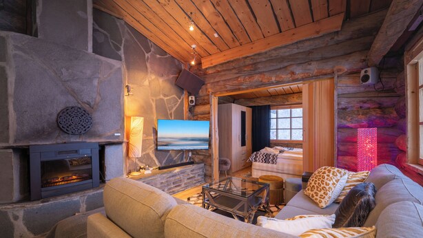 Acogedor interior de cabaña de madera con chimenea de piedra, un sofá cómodo y un televisor.