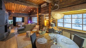 Interior de una cabaña de troncos con una chimenea acogedora, vigas de madera en el techo, una zona de comedor con una mesa dispuesta para cuatro personas y vistas a un paisaje nevado a través de amplios ventanales.