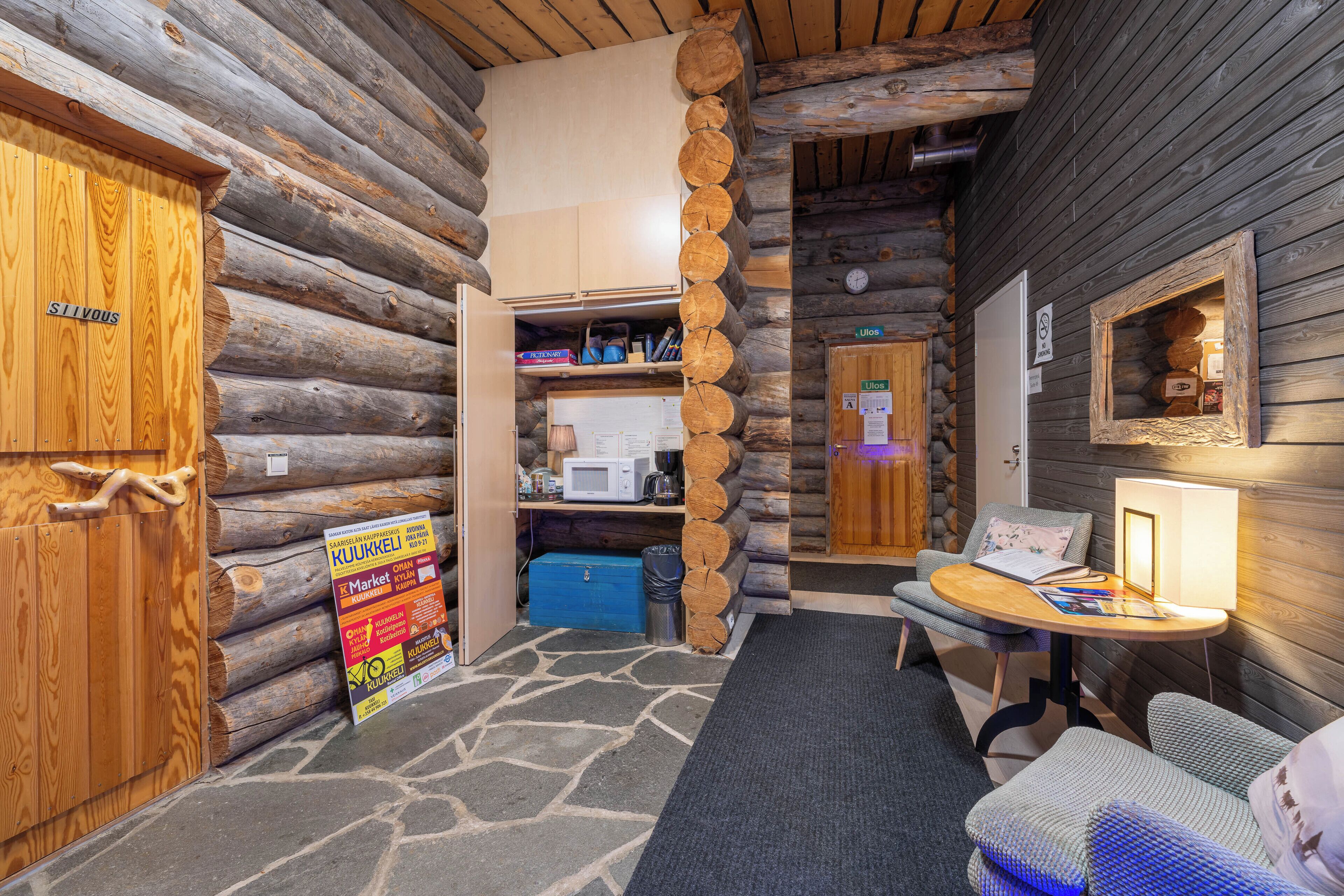 Interior de uma cabana de madeira com soalho de pedra, uma cozinha compacta, uma porta de madeira com a inscrição 'STIVOUS' e uma pequena mesa com uma luminária.