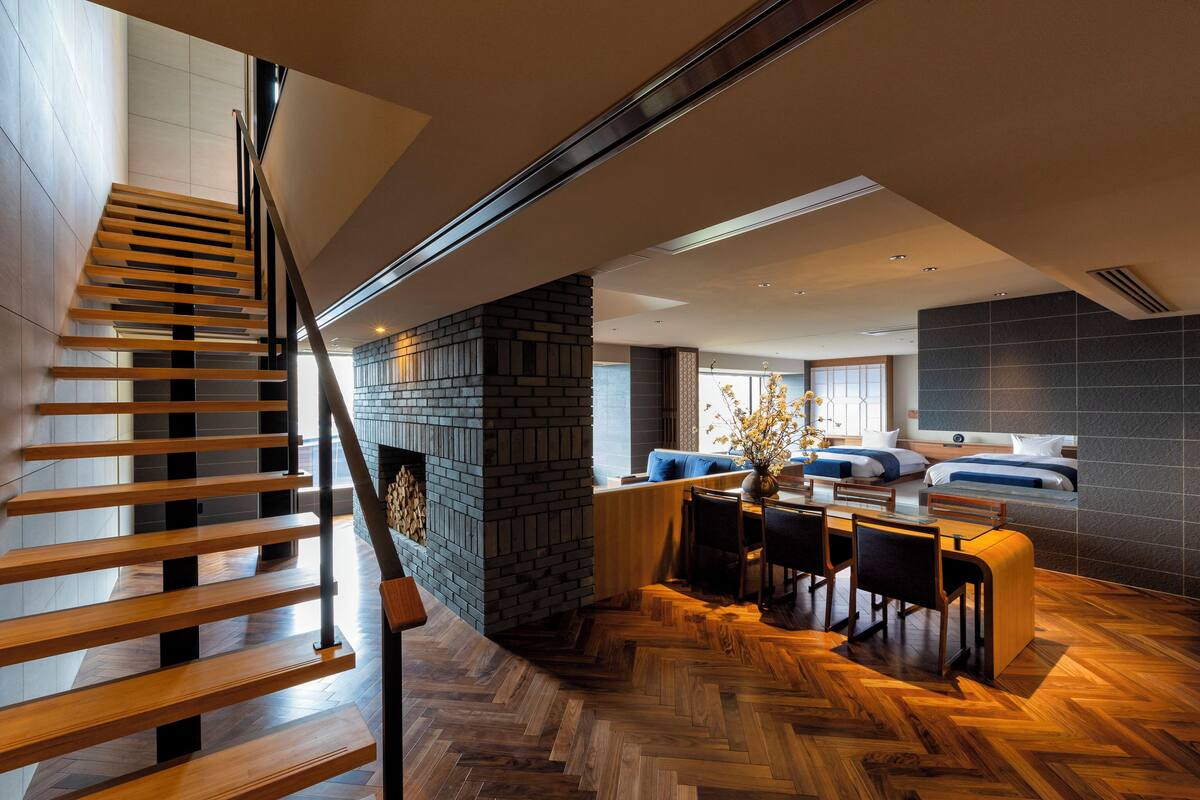 A modern interior with a wooden staircase, a dining area with a table and chairs, and a bedroom with a bed and headboard.