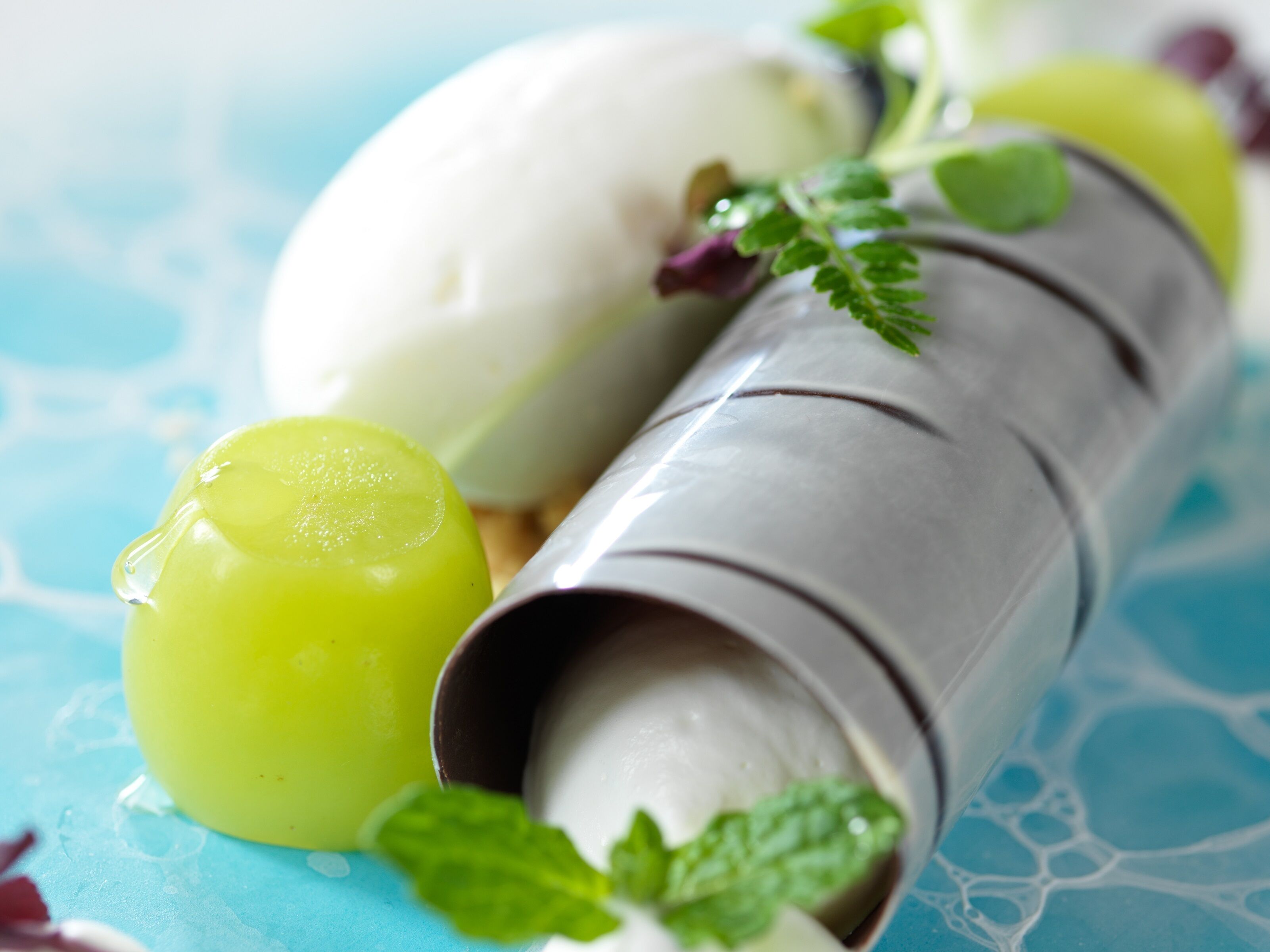A dessert consisting of a scoop of ice cream and a green fruit, possibly a grape, on a blue surface.
