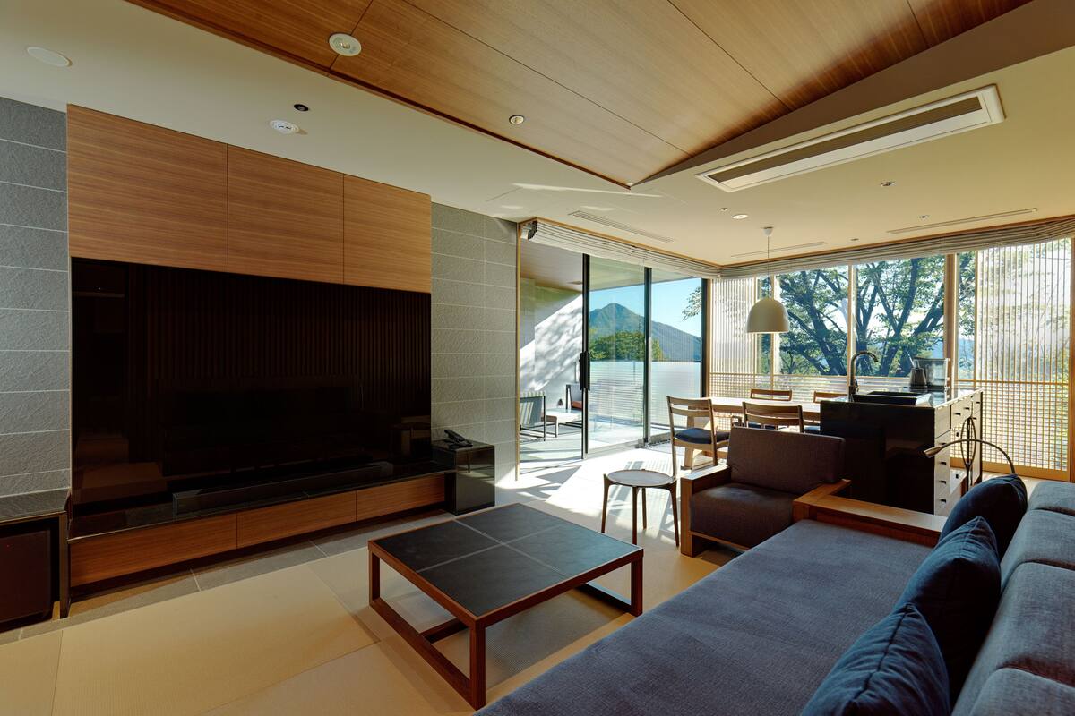 A modern living room with a large flat-screen TV, wooden furniture, and a view of mountains through large windows.