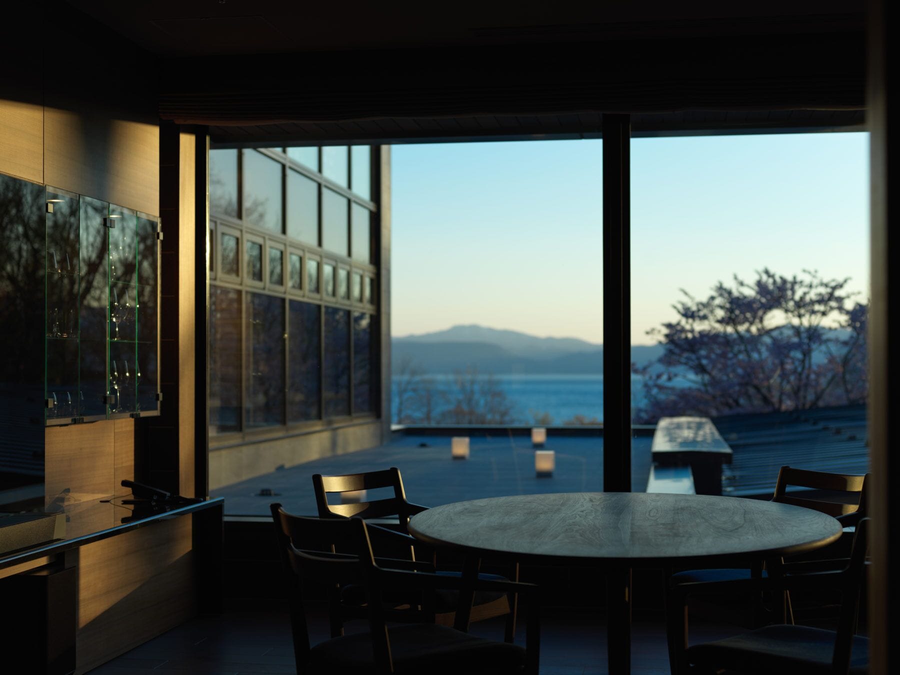 Ein moderner Essbereich mit einem runden Tisch und Stühlen, großen Fenstern mit Blick auf das Meer und die Berge sowie einer Vitrine mit Ausgestellten Gegenständen.