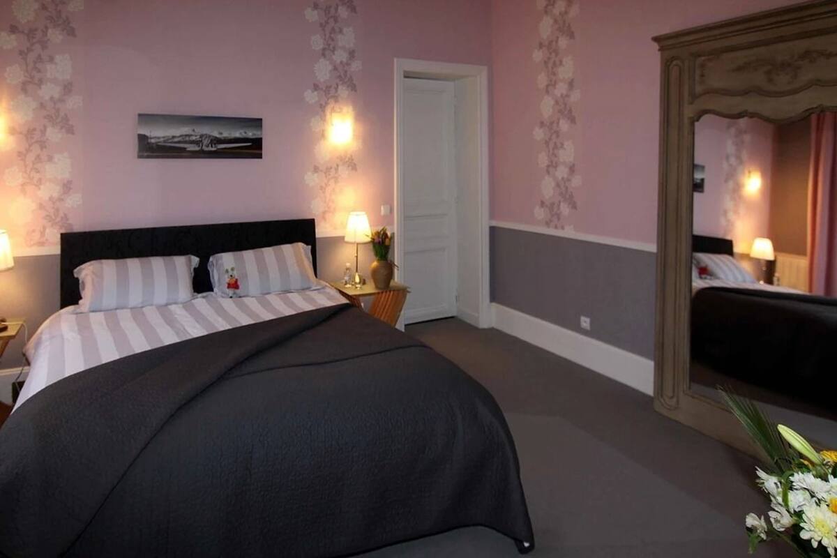 A bedroom with a large bed, a nightstand with a lamp, a dresser, and a mirror.