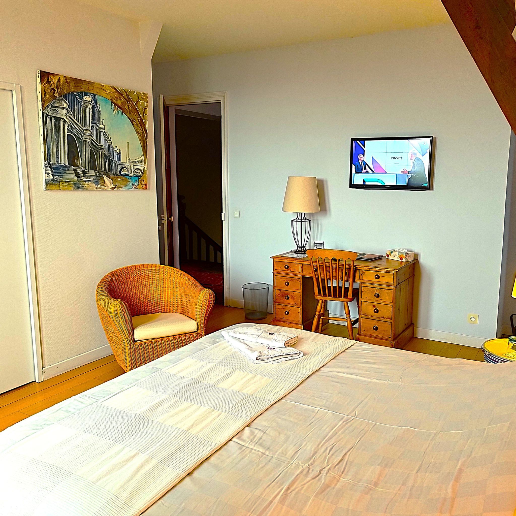 Een slaapkamer met een bed, een stoel, een bureau, een televisie en een schilderij aan de muur.
