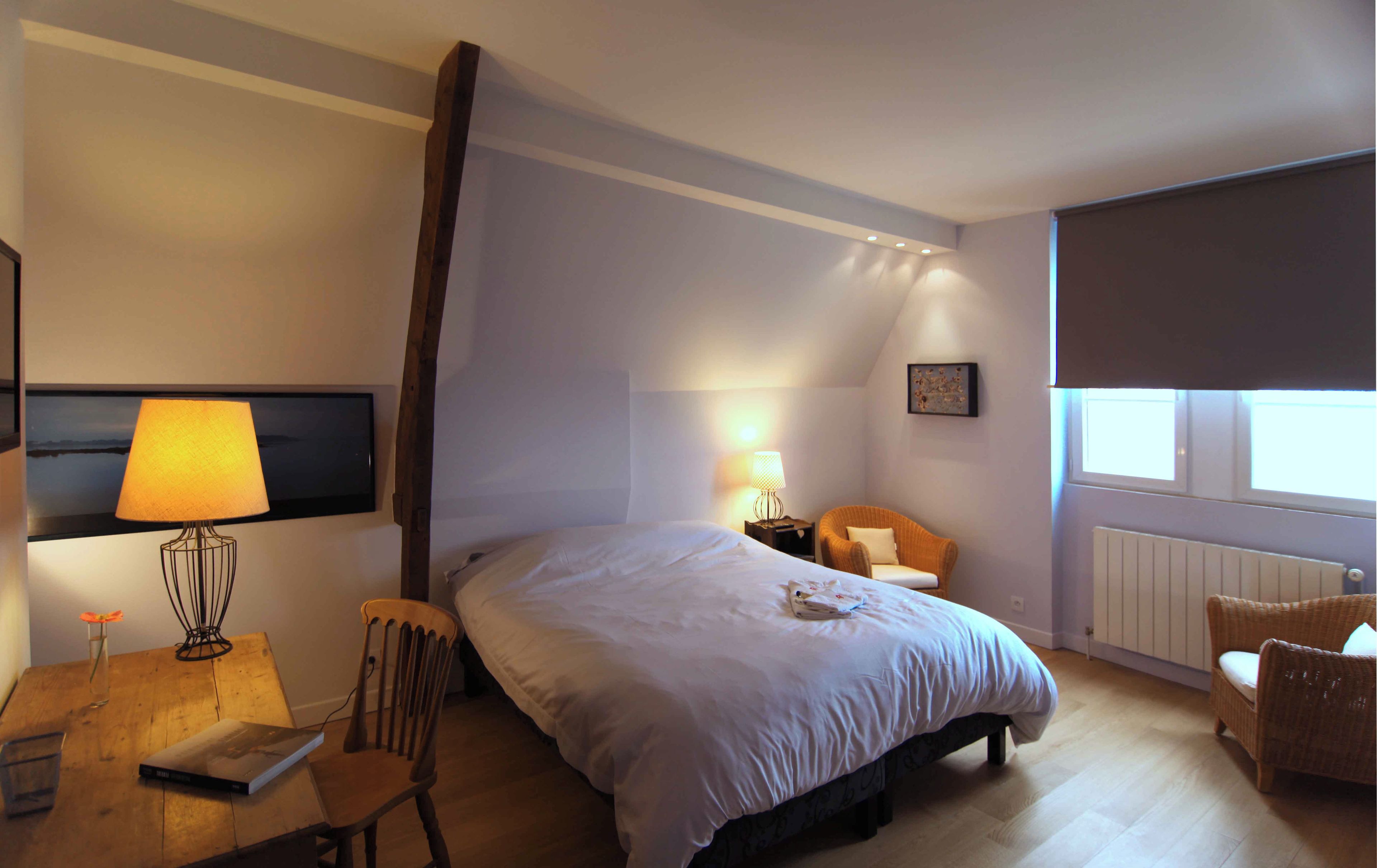 A hotel room with a large bed, a chair, a table with a lamp, and a window.