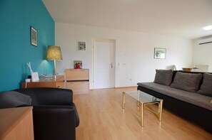A modern living room with a sofa, a coffee table, and a lamp. There is a door leading to another room and a small table with a lamp. - Kliska Apartment (Rovinj)