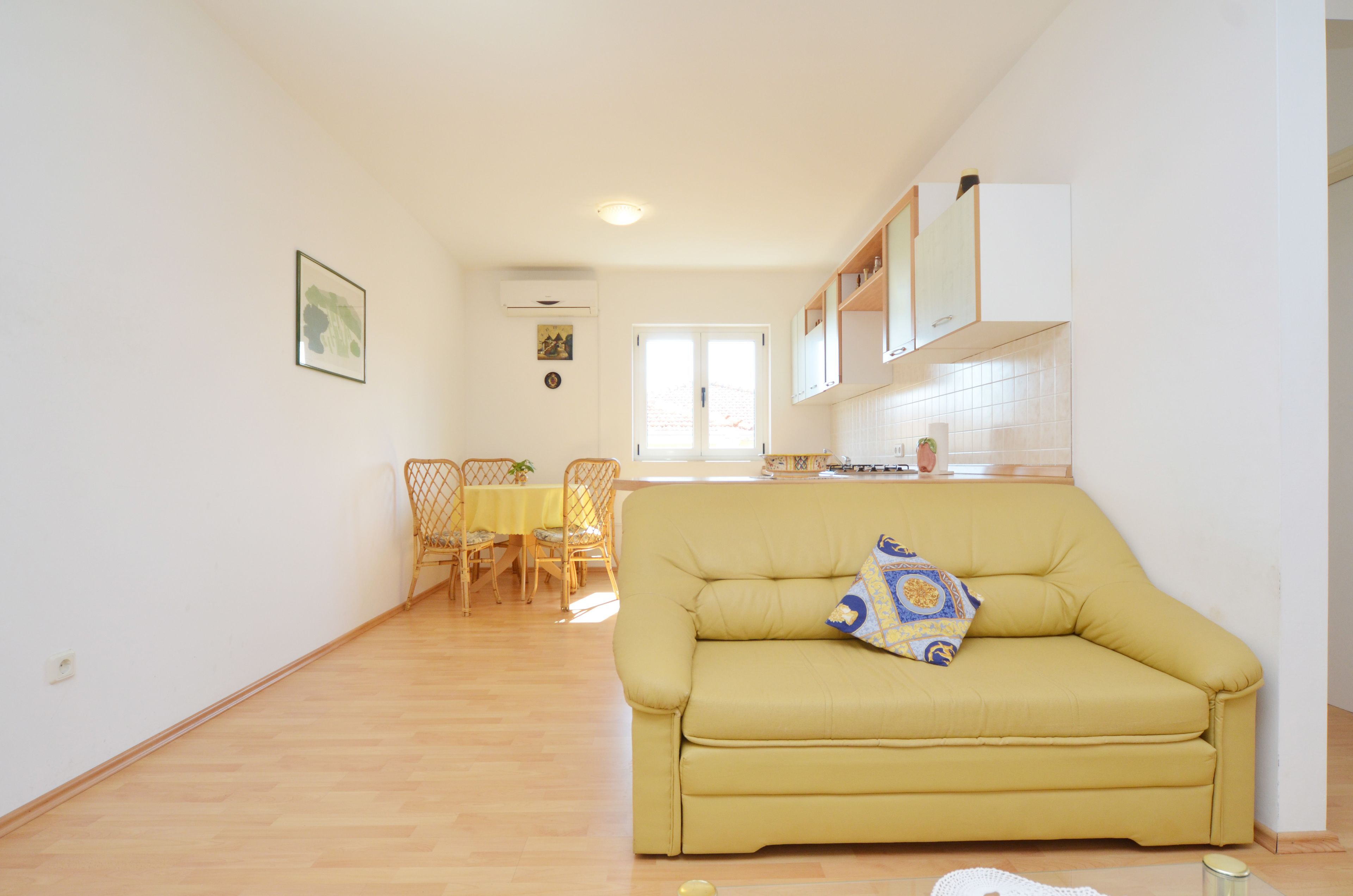 A yellow sofa with a decorative pillow in a bright living room with wooden flooring, a dining area with a table and chairs, and a kitchen with white cabinets and a tiled backsplash.