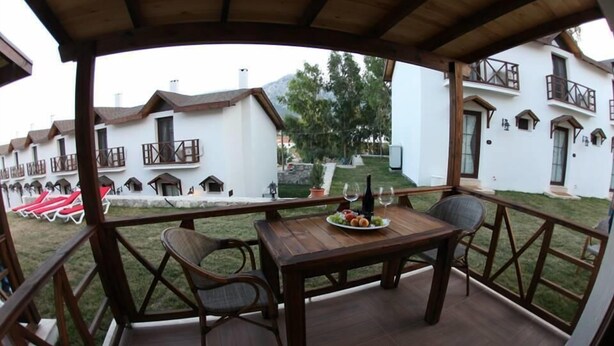 A wooden deck with a table set for two, surrounded by chairs. A plate of fruit and a bottle of wine are on the table. The deck overlooks a garden with a white building in the background. - Karaburun Mimoza Apart Hotel (Karaburun)