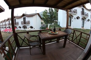 Una terraza de madera con una mesa dispuesta para dos personas, rodeada de sillas. En la mesa hay una fuente de fruta y una botella de vino. La terraza da a un jardín con un edificio blanco al fondo.