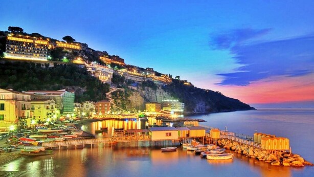 Une ville côtière au crépuscule, avec des bâtiments le long du front de mer, un quai où sont amarrés des bateaux et une mer calme.