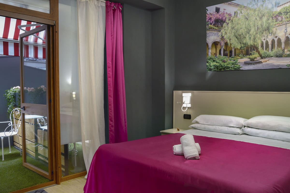 Une chambre d’hôtel comprenant un lit, une table de chevet, une lampe fixée au mur, une fenêtre avec des rideaux et une vue sur un patio extérieur.
