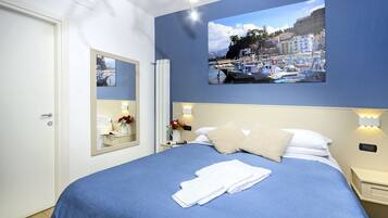 Habitación de hotel con cama, mesita de noche, espejo e imagen en la pared de un pueblo costero.