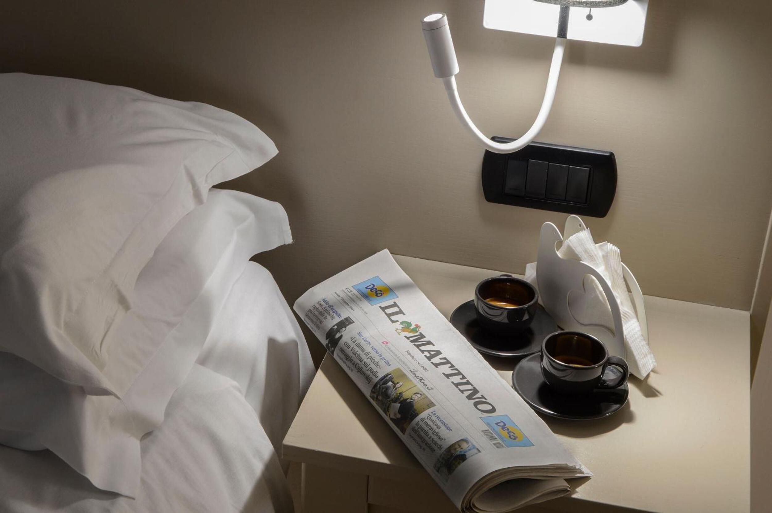 Un lit bien fait, avec des draps blancs, un journal roulé, deux tasses de café et un oreiller blanc posé sur une table de chevet.