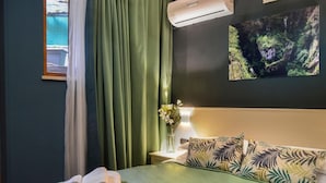 Un lit bien fait, avec une couvre-lit verte et des coussins ornés de motifs de feuilles tropicales, un vase rempli de fleurs sur une table de chevet, et un climatiseur fixé au mur.