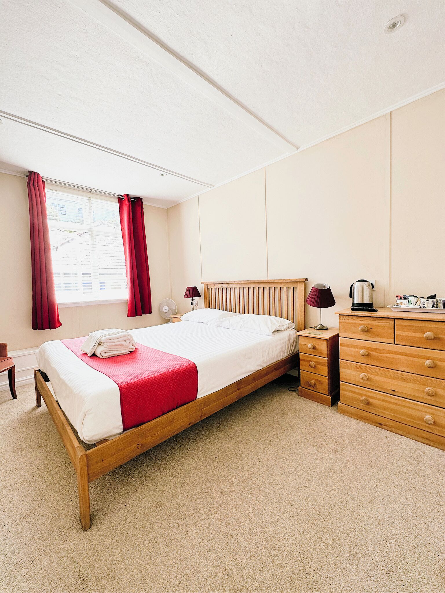 A bedroom with a single bed, a wooden dresser, a nightstand, a lamp, and a window with curtains.