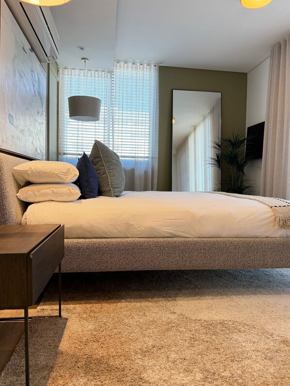 Un dormitorio moderno con una cama grande, una mesita de noche, una lámpara y un ventanal con cortinas.