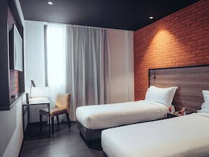 Ein Hotelzimmer mit zwei Betten, einem Schreibtisch, einem Sessel und einem großen Fenster mit Vorhängen.