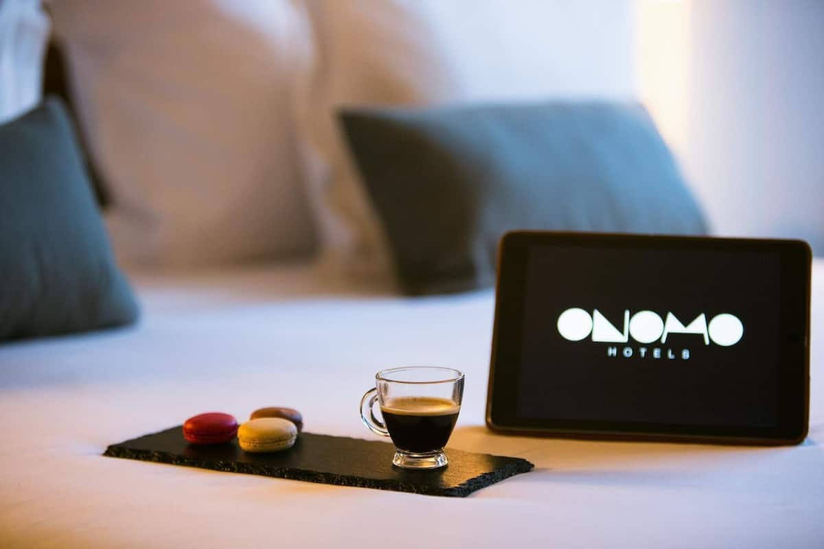 Ein Tablet, das das Logo der ONOMO HOTELS zeigt, auf einem Bett mit einer Kaffeetasse und Macarons.