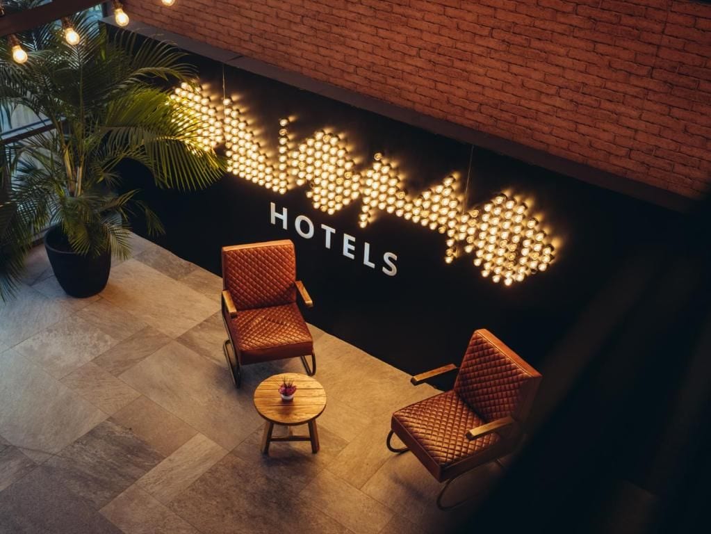 Een hotellobby met een bakstenen muur, een zithoek met twee stoelen en een tafeltje, en een kamerplant.