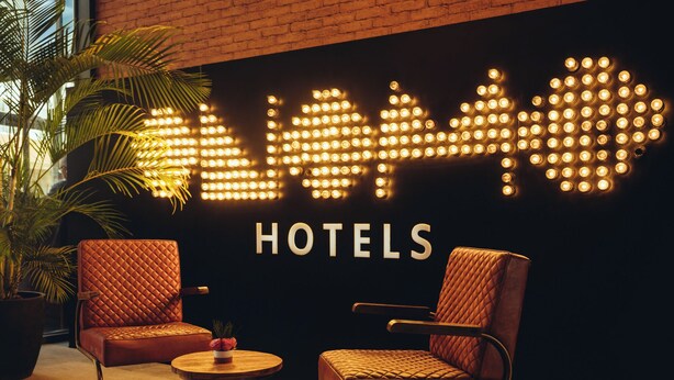 Een hotelschild met verlichte letters en een logo, twee leren fauteuils, een houten tafel en een pot met een plant.