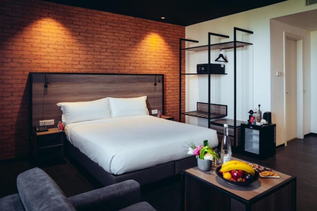 A modern hotel room with a brick accent wall, a large bed, a bedside table with a fruit bowl, and a small desk with a chair.