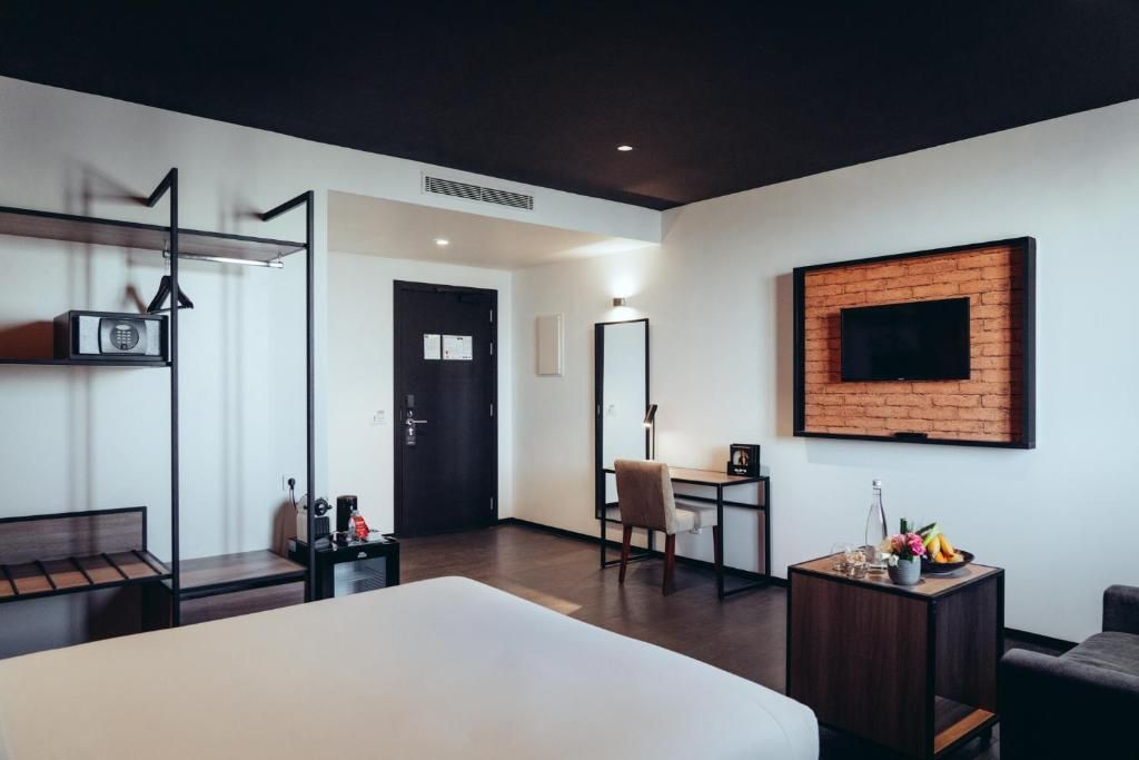 A hotel room with a bed, a desk, a chair, a TV mounted on a brick wall, and a small table with a bottle and fruit.
