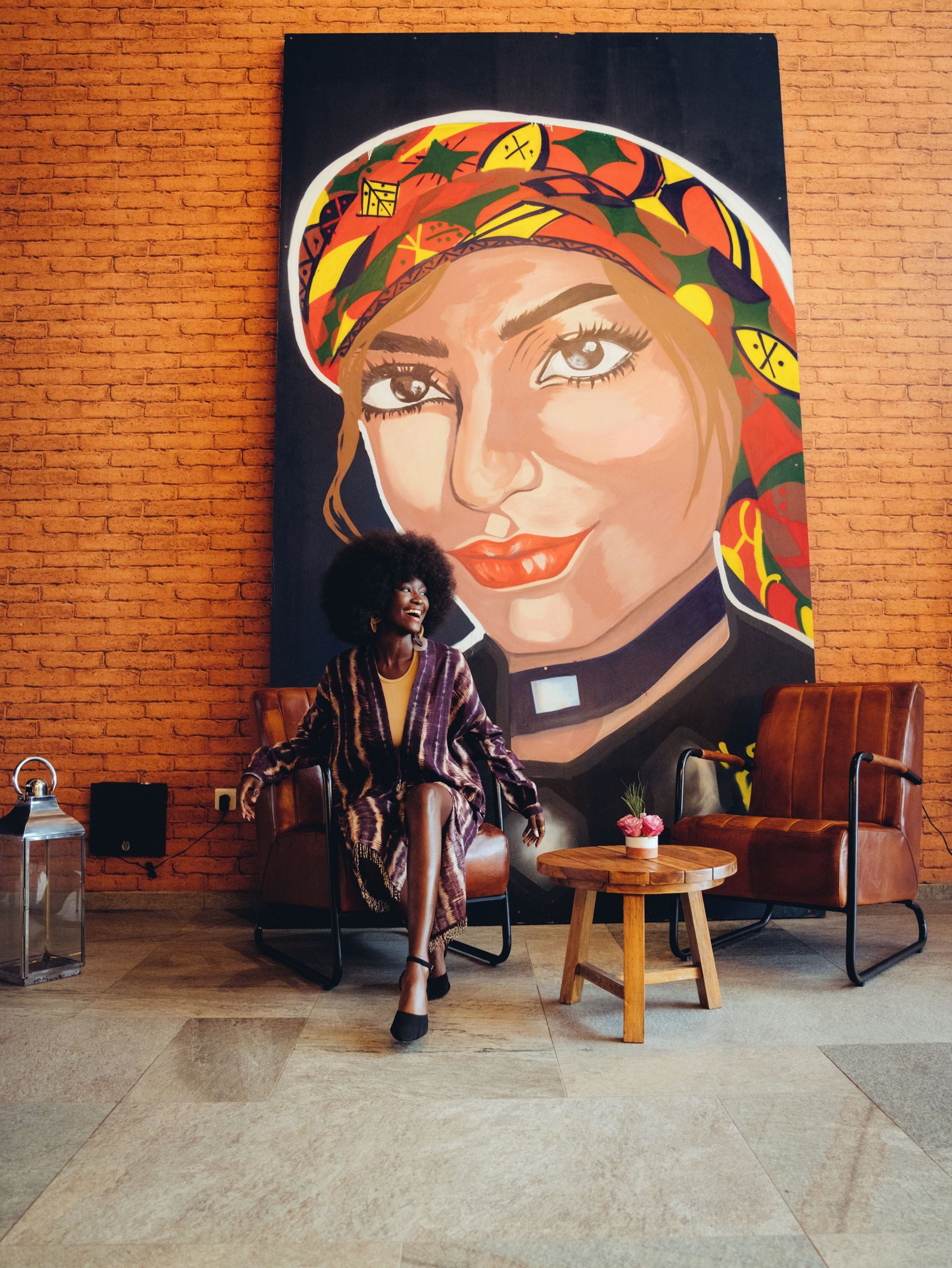 A person seated in a chair in front of a large, colorful mural on a brick wall.
