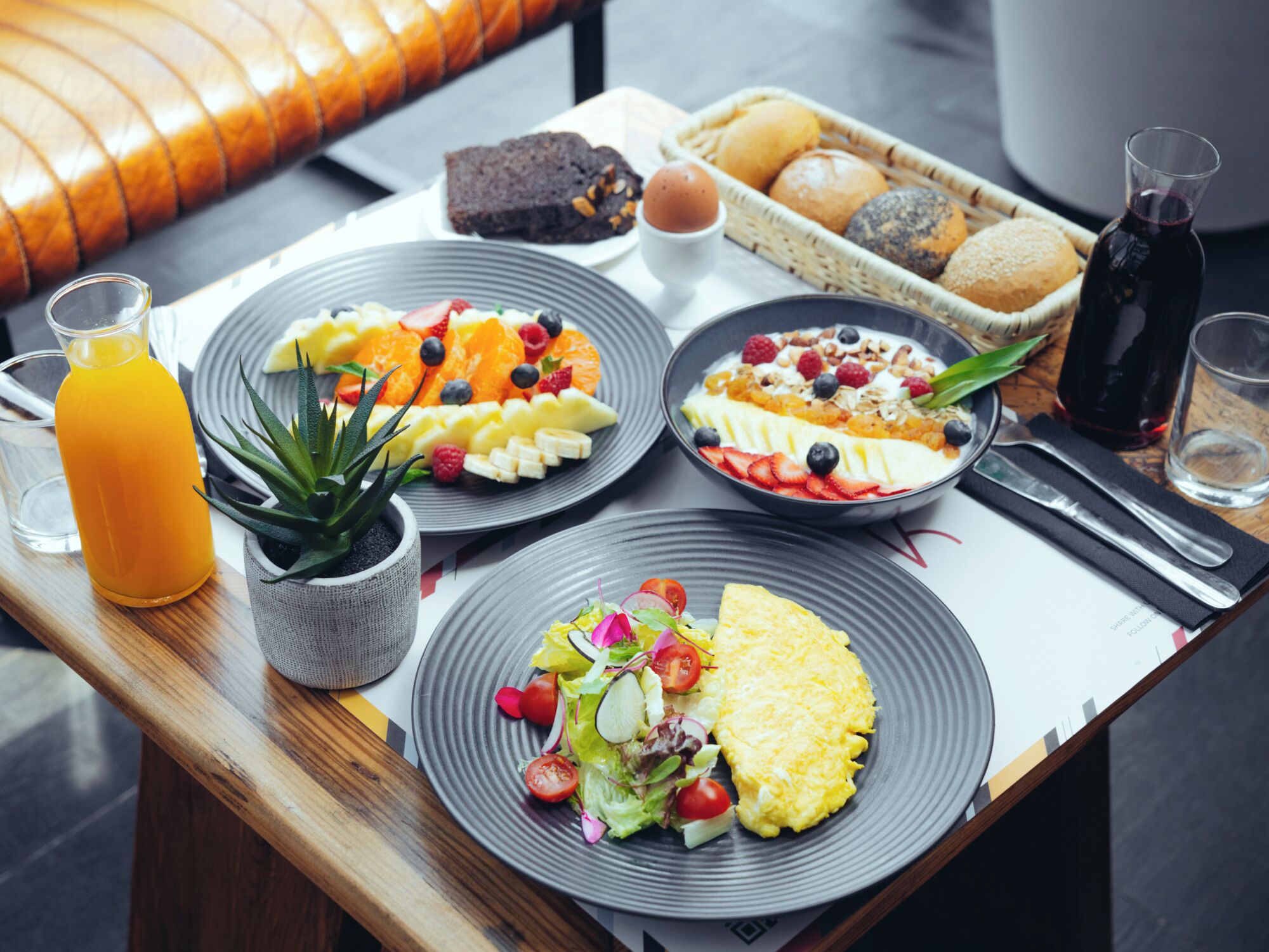 A breakfast table with plates of fruit, a bowl of cereal, a glass of orange juice, and a bottle of wine.