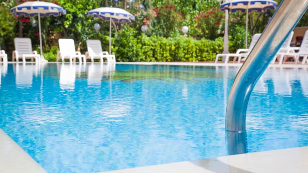 Una piscina con agua azul cristalina, rodeada de sillas reclinables blancas y sombrillas azules.