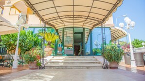 Un espacio al aire libre cubierto con escalones que conducen a la entrada de un edificio, rodeado de plantas en macetas y mobiliario exterior.