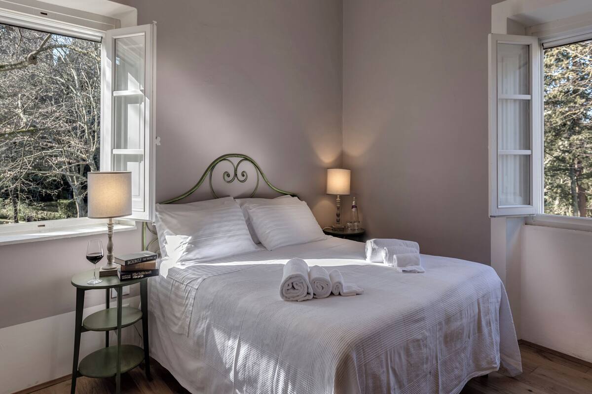 A bedroom with a bed, two bedside lamps, a small table with a book and a glass, and a window with a view of trees.