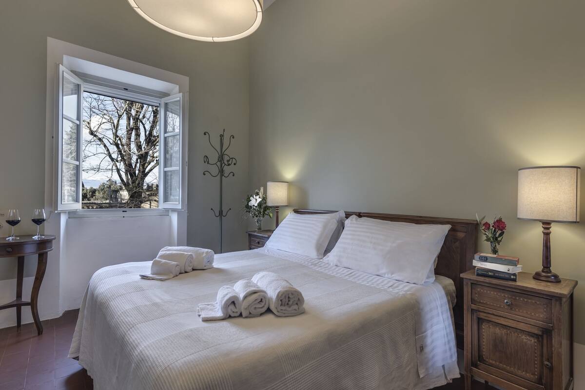 A neatly made bed with white linens and rolled towels, a bedside table with a lamp, and a window with a view of trees.