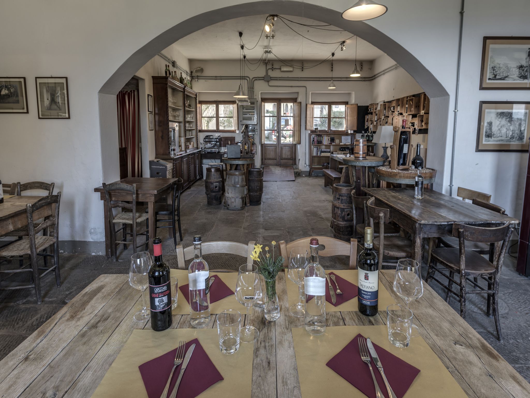 Zona pranzo rustica con tavoli e sedie in legno, bottiglie di vino e bicchieri pronti per un pasto.